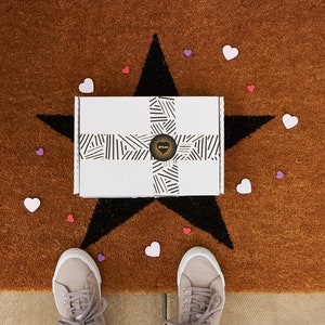 May include: A white box with black and white stripes and a gold heart logo on a brown doormat with a black star and pink and purple hearts. The box is sitting on the star. Two pairs of shoes are on the doormat.