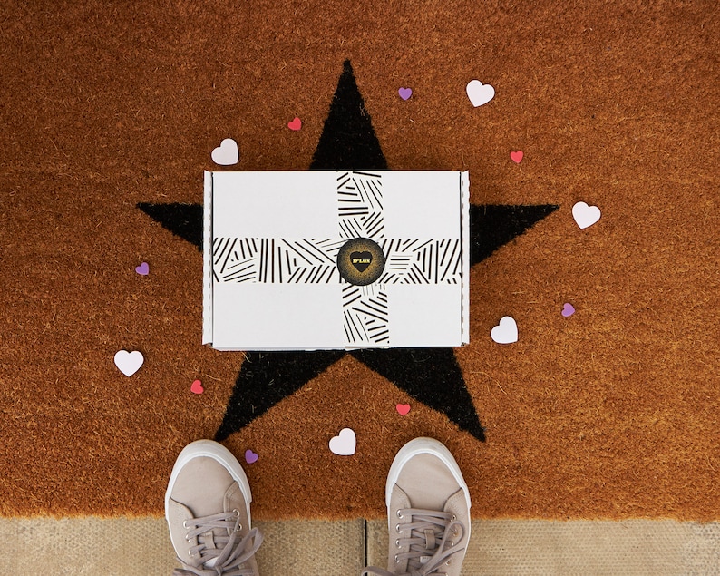 May include: A white box with black and white stripes and a gold heart logo on a brown doormat with a black star and pink and purple hearts. The box is sitting on the star. Two pairs of shoes are on the doormat.
