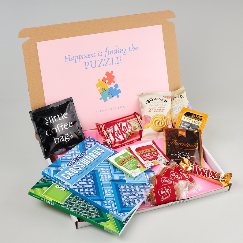K&ouml;nnte beinhalten: Eine braune Geschenkbox mit einem rosafarbenen Deckel, auf dem "Happiness is finding the PUZZLE" steht und ein blaues und gelbes Puzzle-Motiv zu sehen ist. Die Box ist gef&uuml;llt mit verschiedenen Artikeln, darunter eine T&uuml;te Kaffee, ein Kreuzwortr&auml;tselbuch, eine Schachtel Schokolade, eine Schachtel Tee und eine Schachtel Kekse.