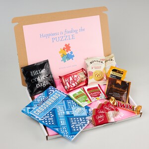 K&ouml;nnte beinhalten: Eine braune Geschenkbox mit einem rosafarbenen Deckel und dem Text "Happiness is finding the PUZZLE" auf dem Deckel. Die Box ist gef&uuml;llt mit verschiedenen Artikeln, darunter ein Kreuzwortr&auml;tselbuch, eine T&uuml;te Kaffee, Schokoladentafeln, Teebeutel, Kakaopulver und Kekse.
