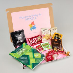 K&ouml;nnte beinhalten: Eine rosa und braune Geschenkbox gef&uuml;llt mit verschiedenen Artikeln, darunter ein Sudoku-Puzzlebuch, Schokoladentafeln, Teebeutel, Kaffeep&auml;ckchen und Kekse. Die Box tr&auml;gt den Text "Happiness is finding the PUZZLE" auf dem Deckel.