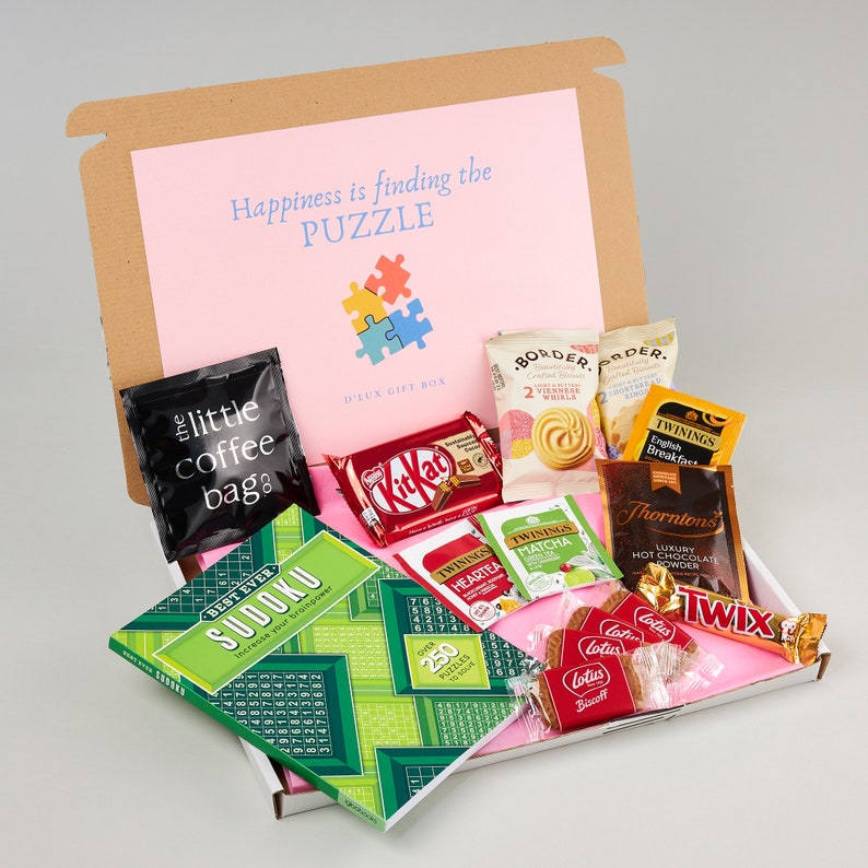K&ouml;nnte beinhalten: Eine rosa und braune Geschenkbox gef&uuml;llt mit verschiedenen Artikeln, darunter ein Sudoku-Puzzlebuch, Schokoladentafeln, Teebeutel, Kaffeep&auml;ckchen und Kekse. Die Box tr&auml;gt den Text "Happiness is finding the PUZZLE" auf dem Deckel.