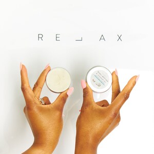 May include: Two hands hold small round containers of shea butter and coconut oil. The containers are white with black text that reads "Shea Butter & Coconut Oil". The word "RELAX" is written in black letters on a white background.