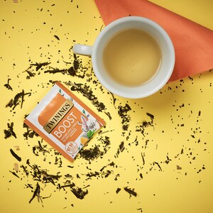 K&ouml;nnte beinhalten: Ein gelber Hintergrund mit einer wei&szlig;en Teetasse gef&uuml;llt mit hellgelbem Tee. Ein einzelner Teebeutel liegt auf der gelben Oberfl&auml;che mit dem Text "TWININGS BOOST" und "Ginger, Pear & Green Tea with Ginseng". Verstreute gr&uuml;ne Teebl&auml;tter liegen um die Teetasse und den Teebeutel herum.