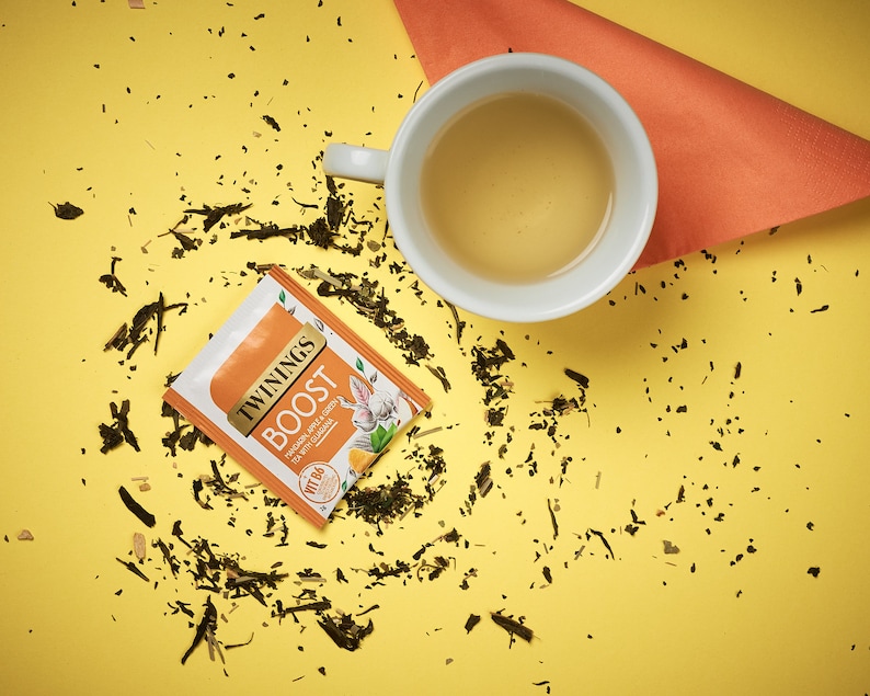 K&ouml;nnte beinhalten: Ein gelber Hintergrund mit einer wei&szlig;en Teetasse gef&uuml;llt mit hellgelbem Tee. Ein einzelner Teebeutel liegt auf der gelben Oberfl&auml;che mit dem Text "TWININGS BOOST" und "Ginger, Pear & Green Tea with Ginseng". Verstreute gr&uuml;ne Teebl&auml;tter liegen um die Teetasse und den Teebeutel herum.