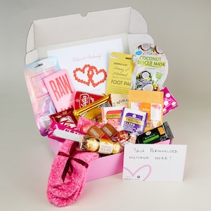May include: Pink gift box filled with various self-care items, including bath products, tea, chocolate, and socks. The box has a pink ribbon and the text "Relax & Recharge!" on the lid. The box also includes a personalized message card.