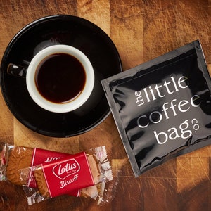 K&ouml;nnte beinhalten: Ein schwarzer Kaffeepackung mit dem Text "the little coffee bag co." darauf gedruckt, eine Tasse Kaffee und zwei einzeln verpackte Lotus Biscoff Kekse auf einer Holzoberfl&auml;che.