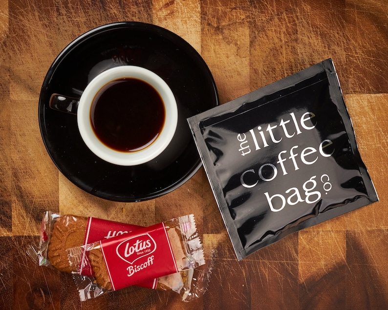 K&ouml;nnte beinhalten: Ein schwarzer Kaffeepackung mit dem Text "the little coffee bag co." darauf gedruckt, eine Tasse Kaffee und zwei einzeln verpackte Lotus Biscoff Kekse auf einer Holzoberfl&auml;che.