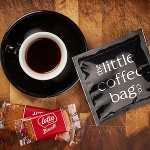 May include: A black coffee bag with the text "the little coffee bag co." printed on it, a cup of coffee, and two individually wrapped Lotus Biscoff cookies on a wooden surface.