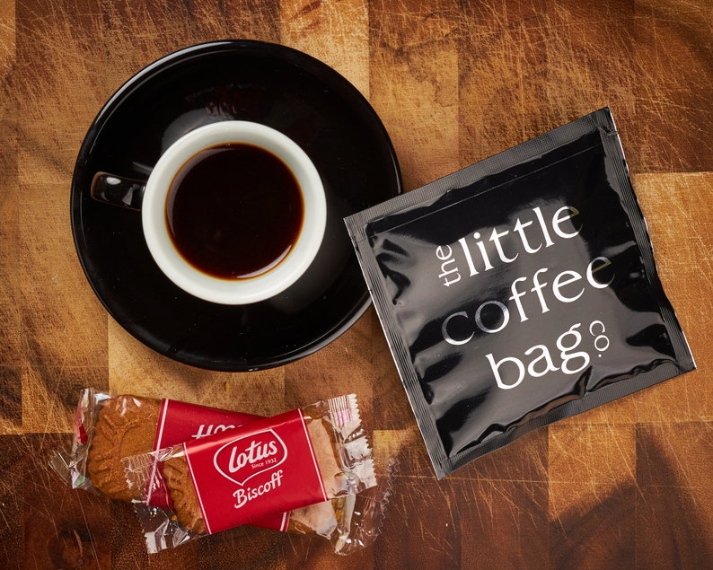 May include: A black coffee bag with the text "the little coffee bag co." printed on it, a cup of coffee, and two individually wrapped Lotus Biscoff cookies on a wooden surface.