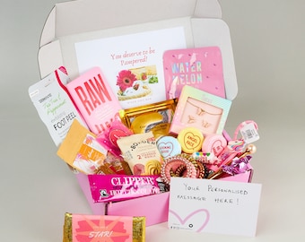 Pode incluir: Uma caixa de presente rosa e branca cheia de vários itens, incluindo produtos de banho, lanches e acessórios. A caixa tem uma mensagem escrita à mão que diz "Sua mensagem personalizada aqui!"
