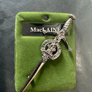 Op de afbeelding: Zilverkleurige kiltpin met een zwaardontwerp en een rond schild met een boom en de tekst "MacLAINE", "VIRTUS" en "MORI", op een groene fluwelen achtergrond.
