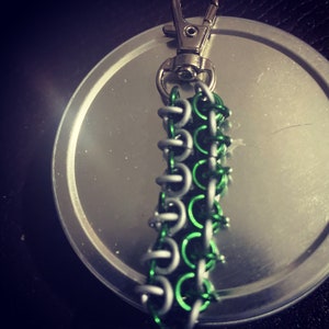 May include: A handmade keychain with a chainmail pattern, using silver and green rings. It has a silver clasp for attachment. The chainmail section is about 7.6 cm long.