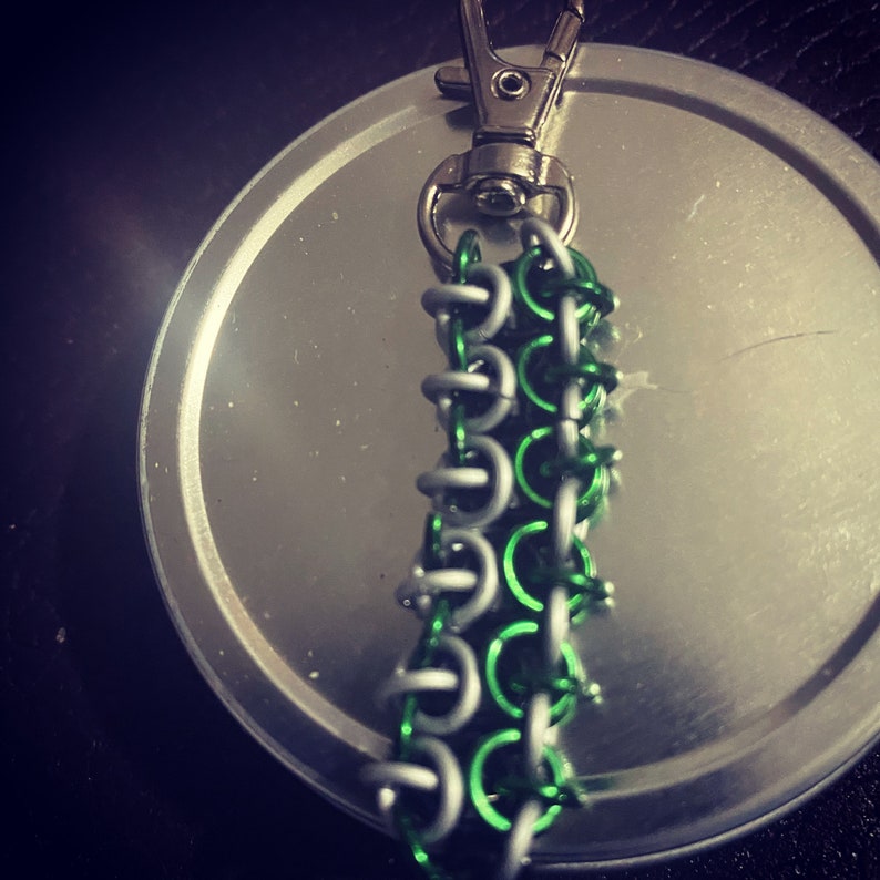 May include: A handmade keychain with a chainmail pattern, using silver and green rings. It has a silver clasp for attachment. The chainmail section is about 7.6 cm long.