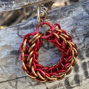 May include: A handmade keychain with a circular design made of interwoven red and gold metal rings. The keychain has a silver-coloured clasp and ring for attaching to keys. The intricate pattern creates a textured appearance.