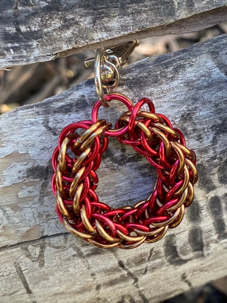 May include: A handmade keychain with a circular design made of interwoven red and gold metal rings. The keychain has a silver-coloured clasp and ring for attaching to keys. The intricate pattern creates a textured appearance.