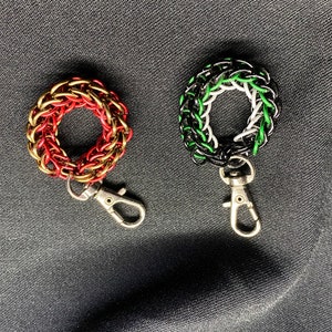 May include: Two chainmaille keyrings. One is red and gold, the other is black, white, and green. Each keyring has a circular design and a silver-coloured clasp. The keyrings are displayed on a dark grey fabric.