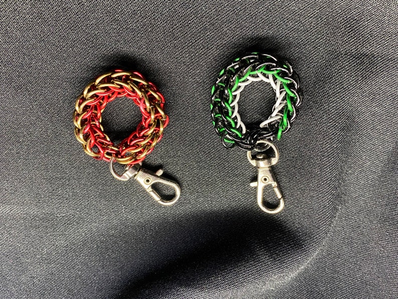 May include: Two chainmaille keyrings. One is red and gold, the other is black, white, and green. Each keyring has a circular design and a silver-coloured clasp. The keyrings are displayed on a dark grey fabric.