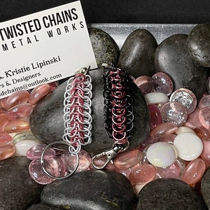 May include: Two chainmail keychains, one silver and red, the other black and red, are displayed on a bed of polished stones. The keychains are made by Twisted Chains Metal Works, as indicated by the business card in the background.