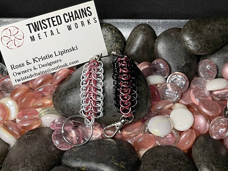 May include: Two chainmail keychains, one silver and red, the other black and red, are displayed on a bed of polished stones. The keychains are made by Twisted Chains Metal Works, as indicated by the business card in the background.