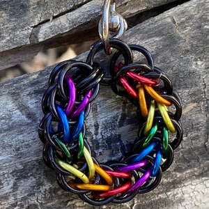 May include: A circular pendant made of black metal, featuring a woven design in rainbow colours. The pendant has a silver clasp. The rainbow includes red, orange, yellow, green, blue, and purple.