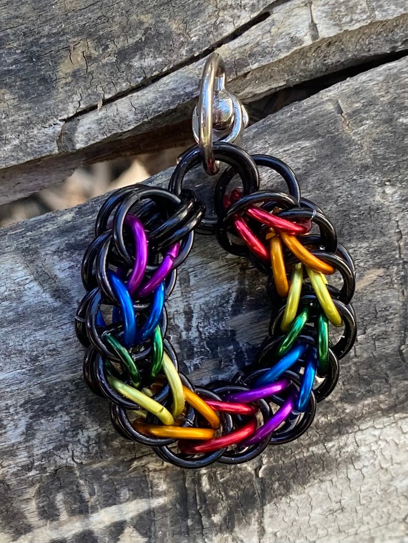 May include: A circular pendant made of black metal, featuring a woven design in rainbow colours. The pendant has a silver clasp. The rainbow includes red, orange, yellow, green, blue, and purple.