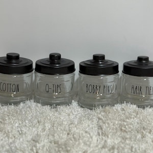 Op de afbeelding: Vier heldere glazen potten met zwarte deksels, met de labels "Cotton", "Q-Tips", "Bobby Pins" en "Hair Ties". De potten staan op een wit, getextureerd oppervlak.