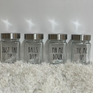 Pode incluir: Quatro frascos de vidro transparente com tampas prateadas, cada um com uma frase preta: "JUST THE TIP", "BALLS DEEP", "PIN ME DOWN" e "TIE ME UP". Os frascos estão sobre uma superfície branca texturizada.