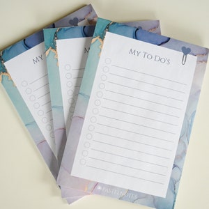 Könnte beinhalten: Ein Stapel aus vier Notizblöcken mit To-Do-Listen in blauem und violettem Marmormuster mit einer silbernen Büroklammer auf dem obersten Notizblock. Der Text "MY TO DO'S" ist auf dem obersten Notizblock gedruckt. Der Text "PASTELNOTES" ist auf dem untersten Notizblock gedruckt.