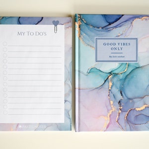 Könnte beinhalten: Ein blaues und violettes Notizbuch mit Marmor-Design und goldenen Akzenten. Der Einband trägt die Aufschrift "Good Vibes Only" und "My little notebook". Das Notizbuch liegt neben einem weißen Notizblock mit einer Checkliste und den Worten "My To Do's".