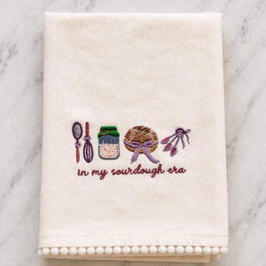 Op de afbeelding: Crèmekleurige keukendoek met geborduurde zuurdesem bakgereedschappen en de zin "in my sourdough era" in paars. De handdoek heeft een pompom-rand langs de onderkant.