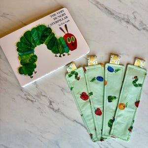 May include: A copy of "The Very Hungry Caterpillar" book by Eric Carle, next to four green fabric bookmarks. The bookmarks feature fruit and caterpillar designs. The book cover shows a green caterpillar with a red head.