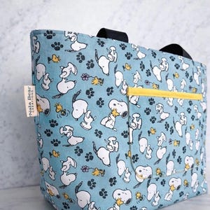 Op de afbeelding: Een lichtblauwe tote bag met een herhaald patroon van Snoopy en Woodstock karakters, pootafdrukken en bloemen. De tas heeft een gele zak en zwarte handvatten. Een label aan de zijkant leest "Nala Bear Creations."