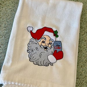 Op de afbeelding: Crèmekleurige kersthanddoek met een geborduurd ontwerp van de kerstman. De kerstman draagt een rode muts en houdt een blikje Diet Coke vast. De handdoek heeft een pomponrand langs de onderkant.