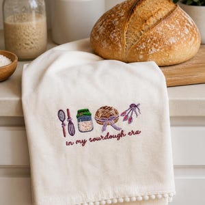 Op de afbeelding: Cr&egrave;mekleurige keukendoek met geborduurde paarse en groene zuurdesem bakpictogrammen en de tekst "in my sourdough era". De handdoek heeft een pompomrand. Een brood ligt in de buurt.