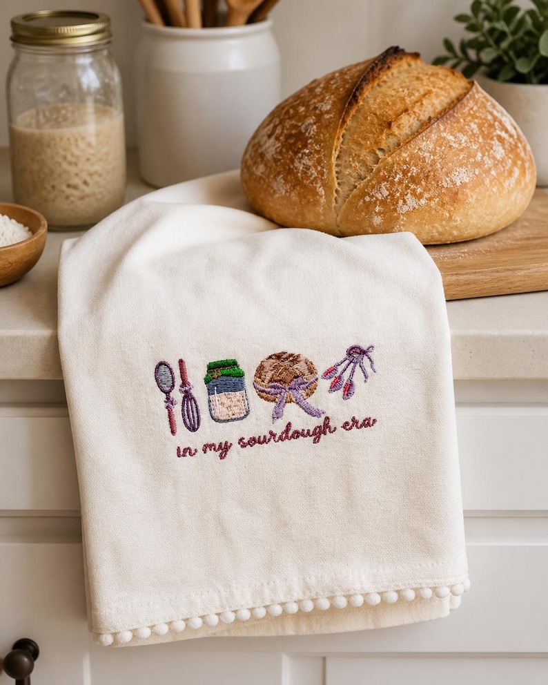 Op de afbeelding: Cr&egrave;mekleurige keukendoek met geborduurde paarse en groene zuurdesem bakpictogrammen en de tekst "in my sourdough era". De handdoek heeft een pompomrand. Een brood ligt in de buurt.