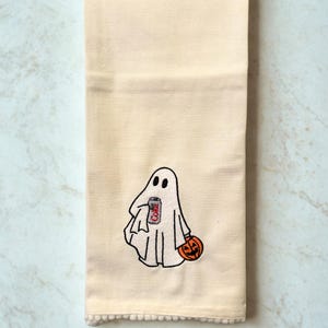 Op de afbeelding: Crèmekleurige Halloween-keukendoek met een geborduurde geest met een frisdrankblikje en een uitgesneden pompoen. De geest is wit met zwarte ogen. Het frisdrankblikje is rood en wit. De onderkant heeft een crèmekleurige pompom-afwerking.