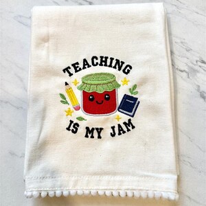 Op de afbeelding: Crèmekleurige keukendoek met geborduurd ontwerp. Het heeft een lachende rode jam pot, een gele potlood, een blauw boek en de tekst "TEACHING IS MY JAM". De onderkant is afgewerkt met een rij kleine witte pompons.