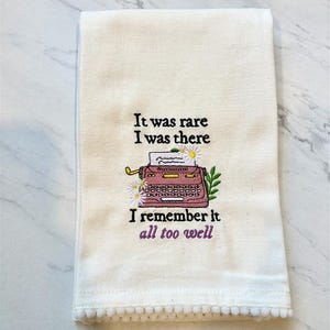 Op de afbeelding: Crèmekleurige keukendoek met geborduurde tekst en een typemachinedesign. De tekst luidt "It was rare I was there I remember it all too well." De typemachine is roze met witte madeliefjes en groene bladeren.