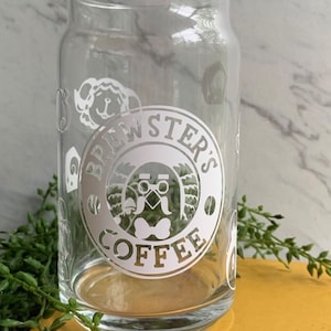 May include: A clear glass mug with a white logo that says "Brewster's Coffee" with a cartoon character wearing glasses and a bow tie. The logo is surrounded by a circle with a coffee bean and a leaf design.