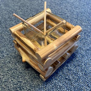 Puede incluir: Un palillero rústico de madera con un inserto de vidrio transparente. El soporte está construido con pequeños troncos, creando una apariencia de cabaña. Dos palillos de madera se colocan dentro del recipiente de vidrio. El diseño es simple y natural.