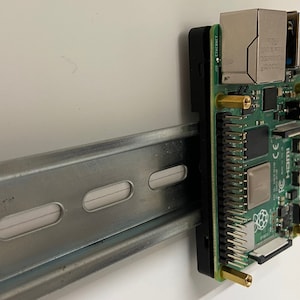 Puede incluir: Una placa de computadora Raspberry Pi verde montada en un riel DIN metálico con una caja negra y tornillos dorados. La placa tiene un puerto de red, una ranura para tarjeta micro SD y un puerto USB. La placa está etiquetada como "Raspberry Pi" y "BCM2837".