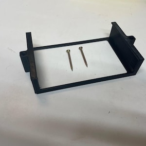 May include: A black plastic frame with two screws. The frame is designed to hold a small object, such as a phone or a tablet.
