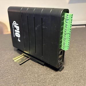 Puede incluir: Un dispositivo electrónico negro con el texto "iPIO-8" en el lateral. Tiene conectores verdes y está montado en un soporte metálico. El dispositivo está sobre una superficie gris.