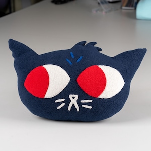 May include: A plush blue cat with red eyes and white pupils. The cat has a simple embroidered face with a nose and whiskers.