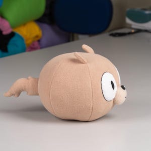 May include: A plush brown toy with a round body and a small tail. The toy has large, white eyes and a black nose.