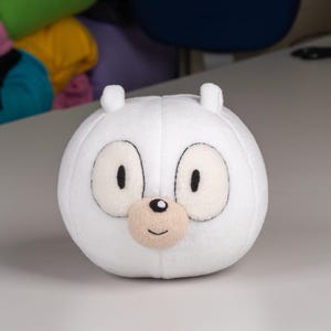 May include: White plush toy with a round shape and a cartoon face. The toy has black eyes, a small black nose, and a pink mouth.