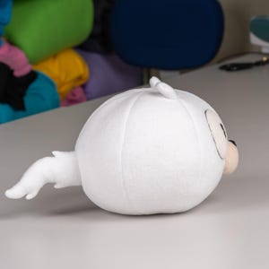 May include: White plush toy shaped like a round, cartoon character with a long, curled tail. The toy has black eyes and a black nose.