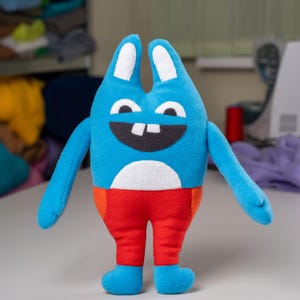 Könnte beinhalten: Ein blaues Plüschtier mit langen Ohren, einem breiten Lächeln und zwei großen weißen Zähnen. Das Spielzeug trägt eine rote Hose und hat einen weißen Bauch.