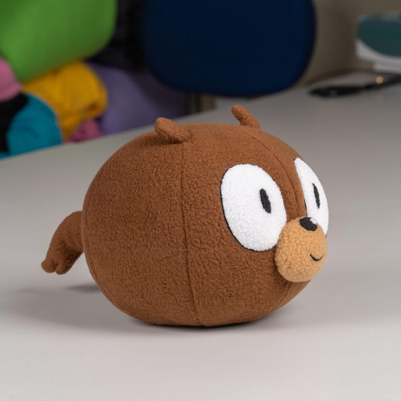 May include: A brown plush toy shaped like a bear with white eyes and a brown nose. The bear has a small tail and is sitting on a white surface.